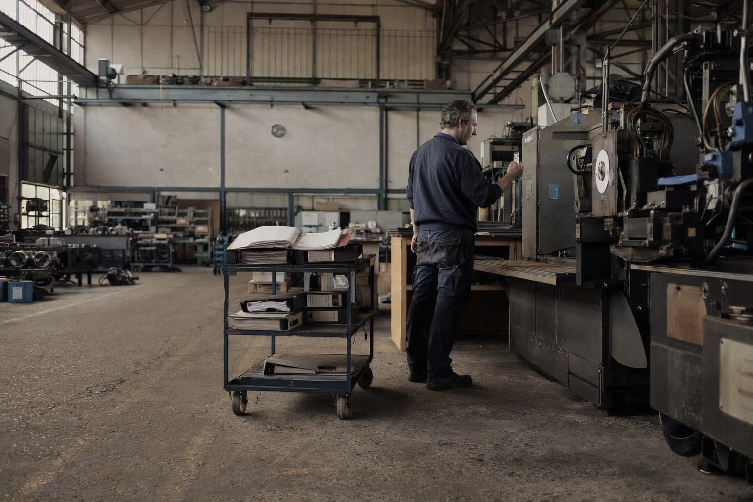 Fitter reading a code off the nameplate of an industrial machine with a smartphone, next to a cart stacked with paper binders
